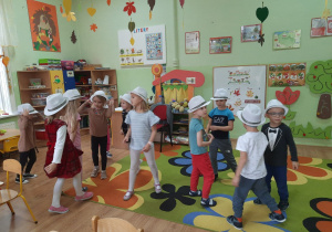 Grupa dzieci w białych kapeluszach na głowach tańczy w rytm muzyki.