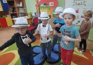 Czterech chłopców w białych kapeluszach na głowach improwizuje gestami grę na gitarach.