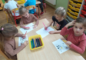 Dziewczynki siedzą przy stoliku i kolorują ilustracje bajkowych postaci.