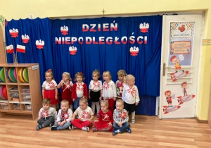 Dzieci z grupy "Słoneczka" w odświętnych strojach na tle granatowej dekoracji z napisem Święto Niepodległości.