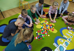 Dzieci na dywanie przeliczają korki w zabawie matematycznej "Ile jabłek ma jeżyk?"