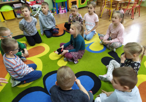 Dzieci siedzą w kole. Pośrodku dziewczynka trzyma w rączkach maskotkę jeża. Dzieci bawią się w zabawę "Na dywanie siedzi jeż"