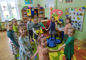 Dzieci tańczą w kole. W środku dziewczynka trzyma w rękach bukiet wiosennych kwiatów.