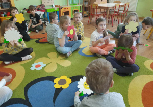 Grupa dzieci siedzi na dywanie z papierowym kwiatkami w rączkach.