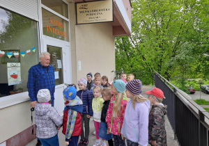 Grupa przedszkolaków stoi przed wejściem do biblioteki i wita się z panem bibliotekarzem.