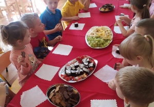 Dzieci siedzą przy stole ze słodko-owocowym poczęstunkiem.