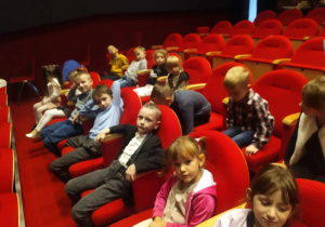 Chłopcy i dziewczynki na widowni na czerwonych fotelach.