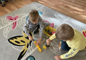Dwóch chłopców na dywanie wkłada małe przedmioty spinaczem do pudełeczka.