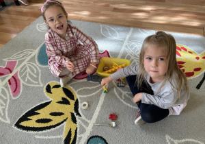 Dwie dziewczynki siedzą na dywanie i wkładają do pudełeczka małe przedmioty za pomocą spinacza.