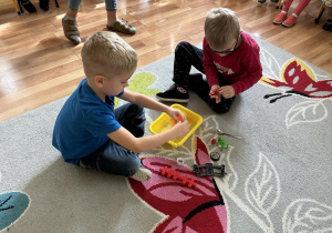 Dwóch chłopców na dywanie wkłada małe przedmioty spinaczem do pudełeczka.