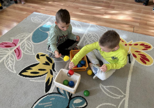 Dwóch chłopców przekłada piłeczki z jednego pudełka do drugiego za pomocą łyżki wazowej.