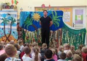 Aktor na tle dekoracji przedstawiającej afrykańską dżunglę zaprasza dzieci na przedstawienie.