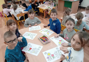 Dzieci przy stoliku kolorują rysunek do programu przedstawiający łąkę z kwiatami i postacią dinozaura Dinusia.