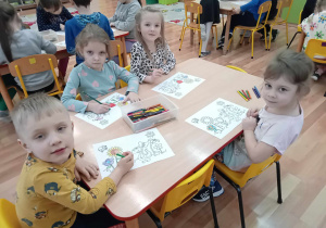 Dzieci przy stoliku kolorują rysunek do programu przedstawiający łąkę z kwiatami i postacią dinozaura Dinusia.