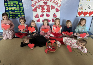 Dziewczynki z grupy Wiewiórek z serduszkami w rękach na tle dekoracji walentynkowej.