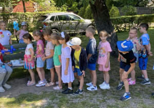 Na ławeczce na ogrodzie przedszkolnym Pan Bartek wykonuje figurki z balonów. Dzieci w rzędzie czekają na swoją kolej.
