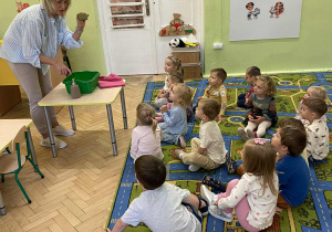 Nauczycielka prezentuje prawidłowy sposób mycia rąk. Na stoliku widzimy miskę z wodą, ręcznik i mydło.