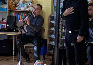 Pan Maciek prowadzący koncert stoi z mikrofonem a obok na krzesełku siedzi i gra na klarnecie współprowadzący koncert.