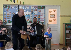 Prowadzący koncert trzyma w rękach koszyk z maskotką węża a obok chłopiec z bębenkiem w rękach, kolorową czapką na głowie gra hipnotyzującą melodię. W tle współprowadzący gra na harmonii.