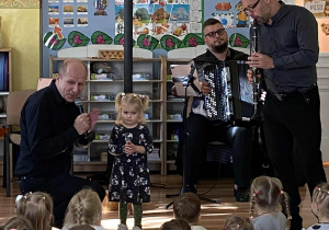 Prowadzący koncert śpiewa do mikrofonu, obok stoi dziewczynka i dwóch muzyków. Jeden gra na harmonii, drugi na klarnecie.