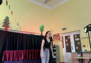 Na zdjęciu widać kobietę stojącą przed grupką małych dzieci w sali. Kobieta ma na sobie czarną bluzkę, jasnoszare spodnie i okulary. Za nią znajduje się scena z zasłoną w kolorze bordowym i czarnym. Dzieci siedzą na podłodze z przodu, zwrócone w stronę kobiety, słuchając jej z zainteresowaniem..