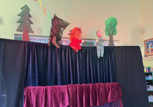 Na zdjęciu widać scenę teatralną przygotowaną do przedstawienia kukiełkowego. Scena jest zasłonięta czarnymi i bordowymi zasłonami, a nad nimi widoczne są trzy kukiełki. Po lewej stronie znajduje się kukiełka wilka o ciemnym kolorze, w środku lalka przedstawiająca dziewczynkę w czerwonej sukience, a po prawej stronie kukiełka chłopca w niebieskim stroju. W tle widoczne są papierowe dekoracje drzewa, które tworzą leśną scenerię.