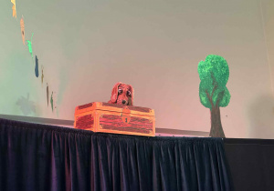 Na górze sceny, za czarną kotarą, stoi skrzynia w kolorze brązowo-złotym z namalowanym zamkiem. Z wnętrza skrzyni wygląda kukiełka psa o brązowej sierści i dużych, wyrazistych oczach. Scena jest oświetlona, co tworzy ciepły, przyjazny klimat.