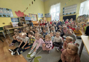 Na zdjęciu widać dużą grupę dzieci siedzących na dywanie w sali przedszkolnej. Dzieci są skupione i z zainteresowaniem oglądają przedstawienie. Sala jest jasno oświetlona, ma żółte ściany ozdobione kolorowymi rysunkami, liśćmi i innymi dekoracjami. W tle widać meble, zabawki, książki oraz plansze edukacyjne z literami i cyframi. Po lewej stronie siedzi nauczycielka, która również obserwuje wydarzenie. Atmosfera jest radosna i pełna zaciekawienia.