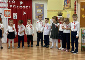 Dzieci w galowych strojach z kotylionami stoją w półkolu deklamując wiersze. W tle dekoracja z napisem Dzień Niepodległości.