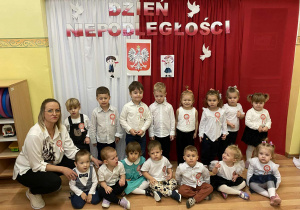 Grupa Wiewiórek pozuje do zdjęcia. W tle dekoracja z napisem Dzień Niepodległości, godłem Polski i ilustracjami dziewczynki i chłopca trzymających w ręce flagę.