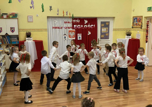 Dzieci ustawione w dwóch kołach tańczą krakowiaka. Chłopcy w wewnętrznym kole trzymają się za ręce, dziewczynki na obwodzie trzymają ręce na biodrach.