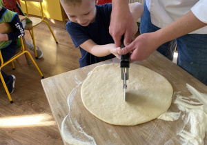 Chłopiec z pomocą właściciela Akademii Pieczenia kroi radełkiem ciasto na rogale.