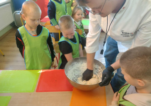 Właściciel Akademii Pieczenia wyrabia w misce ciasto na rogale. Dookoła stoją dzieci.