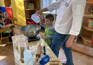 Piekarz z chłopcem przy stole, na którym widzimy mąkę, wagę, miskę, radełko. Mężczyzna wlewa wodę do miski.