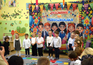 Na zdjęciu widać grupę małych dzieci stojących w szeregu podczas uroczystości w przedszkolu. Są odświętnie ubrane, uśmiechnięte i wyglądają, jakby brały udział w krótkim występie. Za nimi znajduje się duża, bardzo kolorowa dekoracja z napisem Pasowanie na przedszkolaka, przedstawiająca rysunkowe postacie dzieci, balony, kwiaty i girlandy. Przed sceną stoją rodzice lub opiekunowie, którzy obserwują i fotografują wydarzenie. Całość tworzy radosną, uroczystą atmosferę.