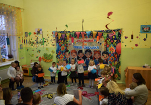 Na zdjęciu widać grupę małych dzieci.. Stoją na środku sali, trzymając w rękach kolorowe kartki, które są elementem ich tańca.