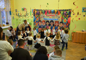 Na zdjęciu widać grupę małych dzieci tańczących w kółku podczas uroczystości Pasowania na Przedszkolaka. Trzymają się za ręce i tworzą duży krąg na kolorowym dywanie w sali przedszkolnej. Na zdjęciu widać także dwie nauczycielki pomagające dzieciom utrzymać koło. Atmosfera jest żywa, pełna entuzjazmu i dziecięcej radości.