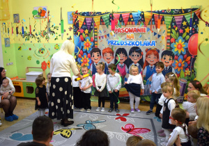 Na środku stoi Pani Dyrektor, która symbolicznym ołówkiem dotyka ramienia jednego z dzieci . Jest to tradycyjny gest pasowania. Całość ma bardzo radosny i uroczysty charakter.