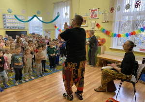 Mężczyzna gra na djembe, a drugi prowadzi rytmiczną zabawę, klaszcząc razem z dziećmi. Przedszkolaki stoją na dywanie i uczestniczą w muzycznych zajęciach.