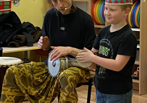 Prowadzący gra na djembe, obok chłopiec w charakterystycznej etnicznej czapce afrykańskiej gra na bębenku.
