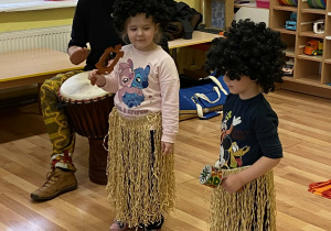 Pan Bartek gra na djembe. Obok niego stoi dwoje dzieci ubrane w złote spódniczki i czarne peruki. W rękach trzymają instrumenty o grają na nich.