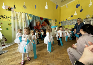 Grupa dzieci stoi w sali przedszkolnej. Dzieci ubrane są na biało. W rękch trzymają jasnoniebieskie szarfy. Przygotowane są do tańca. Za dziećmi powieszone jest tło, na którym przedstawiona jest postać babci i dziadka.