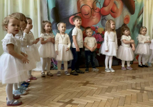 Grupa dzieci ubrana na biało , stoi w rzędzie i śpiewa piosenkę "Jak babcie kocham i dziadka""