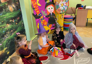 Na zdjęciu widać grupę małych dzieci siedzących na dywanie w przedszkolu. Dzieci są przebrane za czarownice – mają kolorowe kapelusze, peleryny i halloweenowe stroje. W tle znajduje się dekoracja z napisem „Andrzejki” oraz kolorowe ozdoby, co wskazuje na wspólne świętowanie w przedszkolu