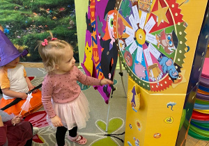 Na zdjęciu dziewczynka w przedszkolu kręci kolorowym kołem fortuny podczas zabawy andrzejkowej, a obok siedzą dzieci w przebraniach.