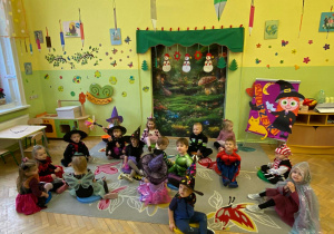 Na zdjęciu grupa dzieci w kolorowych przebraniach siedzi w kręgu na dywanie w przedszkolu podczas zabawy andrzejkowej.