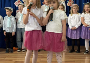 Na pierwszym planie widać dwie dziewczynki mówiące do mikrofonów. Są ubrane w białe bluzki i różowe tiulowe spódniczki. W tle stoją inne dzieci – chłopcy w koszulach z krawatami oraz dziewczynki w fioletowych spódniczkach. Całość odbywa się na tle niebieskiej zasłony ozdobionej papierowymi kwiatami i sercami.