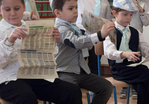 Na zdjęciu widać grupę chłopców siedzących na krzesłach. Chłopcy są ubrani w podobne stroje: kaszkiety, koszule oraz jasnoniebieskie krawaty. Dzieci trzymają w rękach gazetę.