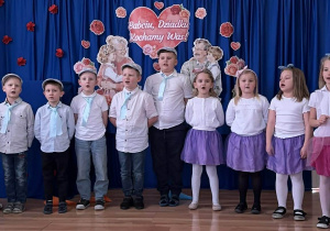 Zdjęcie przedstawia grupę dzieci w wieku przedszkolnym lub wczesnoszkolnym, występujących na scenie z okazji Dnia Babci i Dziadka. Chłopcy ubrani są w białe koszule, niebieskie krawaty oraz kaszkiety. Mają na sobie jeansy lub ciemne spodnie. Dziewczynki mają na sobie białe bluzki i tiulowe spódniczki w kolorze, na głowach mają opaski z kokardkami. Dzieci stoją w rzędzie i śpiewają piosenkę