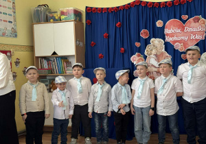 Grupa chłopców w białych koszulach i niebieskich krawatach stoi, gotowa do występu. Tło sceny jest udekorowane niebieską zasłoną, czerwonymi kwiatami i ozdobami w kształcie serc. Centralnym elementem dekoracji jest transparent z napisem "Babciu, Dziadku Kochamy Was!"